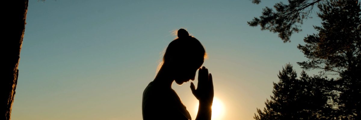 Silhouette of young woman praying in forest at sunset. Sunset light, sun lens flares, golden hour. Religion, spiritual and nature concept